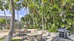 Eine Terrasse im Freien mit Grill, Sitzgelegenheit und üppiger tropischer Bepflanzung.