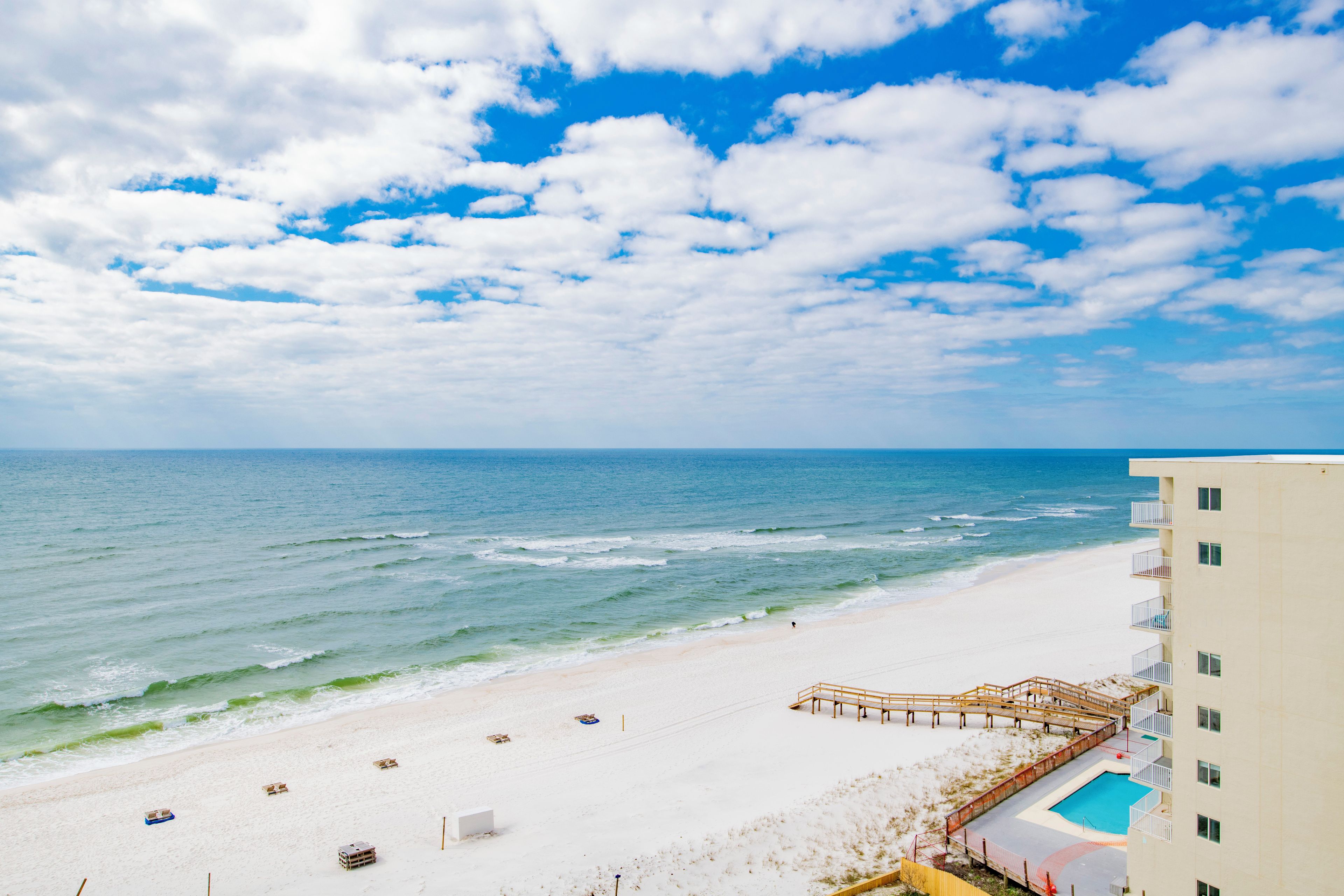 A beach with a wooden walkway leading to a building with a pool.