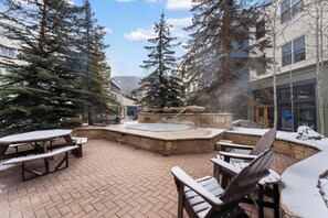 An outdoor patio with a hot tub, wooden chairs, and a stone table. - River Run Condo w/ Shared Hot Tub, Pool & Sauna, Mountain Views from Balcony! (Keystone)
