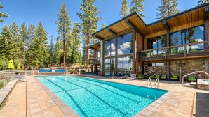 A pool area with a wooden deck, lounge chairs, and a stone building with large windows.