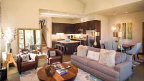 A spacious living room with a large sofa, a coffee table, and a fireplace. There is a kitchen area with dark wooden cabinets and modern appliances.