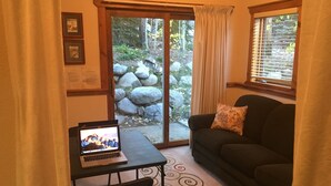 A cozy living room with a laptop on a table, a sofa with a cushion, a sliding glass door leading to a garden with rocks and greenery, and a window with blinds.