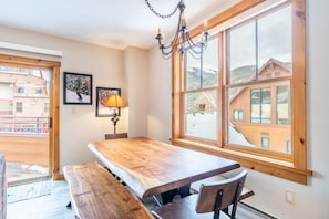 A dining area with a wooden table and benches, large windows with a view of buildings and mountains, and a chandelier.