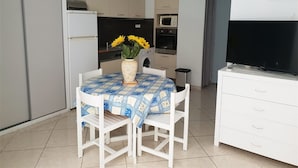Un espacio habitable compacto con comedor que incluye una mesa con mantel a cuadros azules y blancos, dos sillas blancas y un jarrón con flores amarillas. Se ve un mueble blanco con cajones a la derecha, y un televisor de pantalla plana está montado en la pared sobre él.