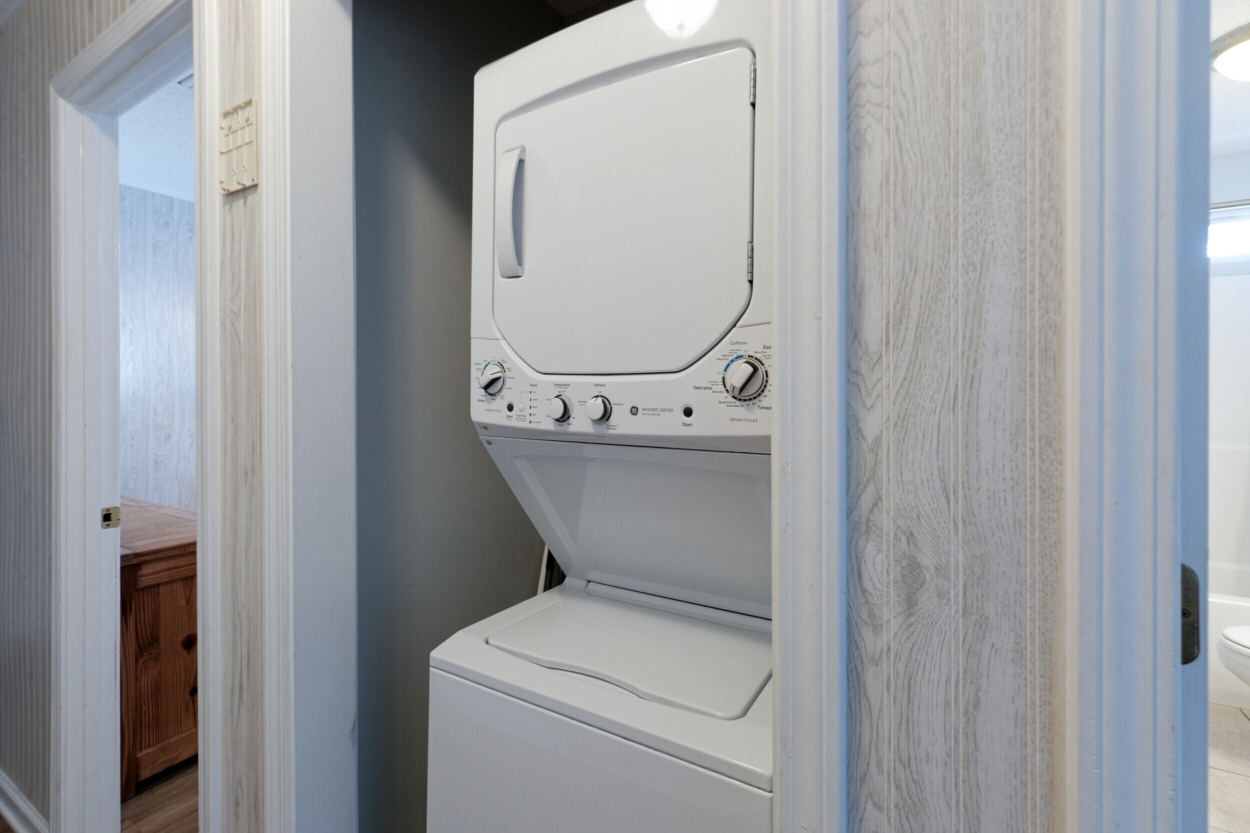 A compact, white washer and dryer unit in a narrow hallway with wooden cabinets and a light blue wall.