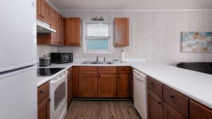 Una cocina con armarios de madera, encimera blanca, estufa, microondas y ventana con persianas.