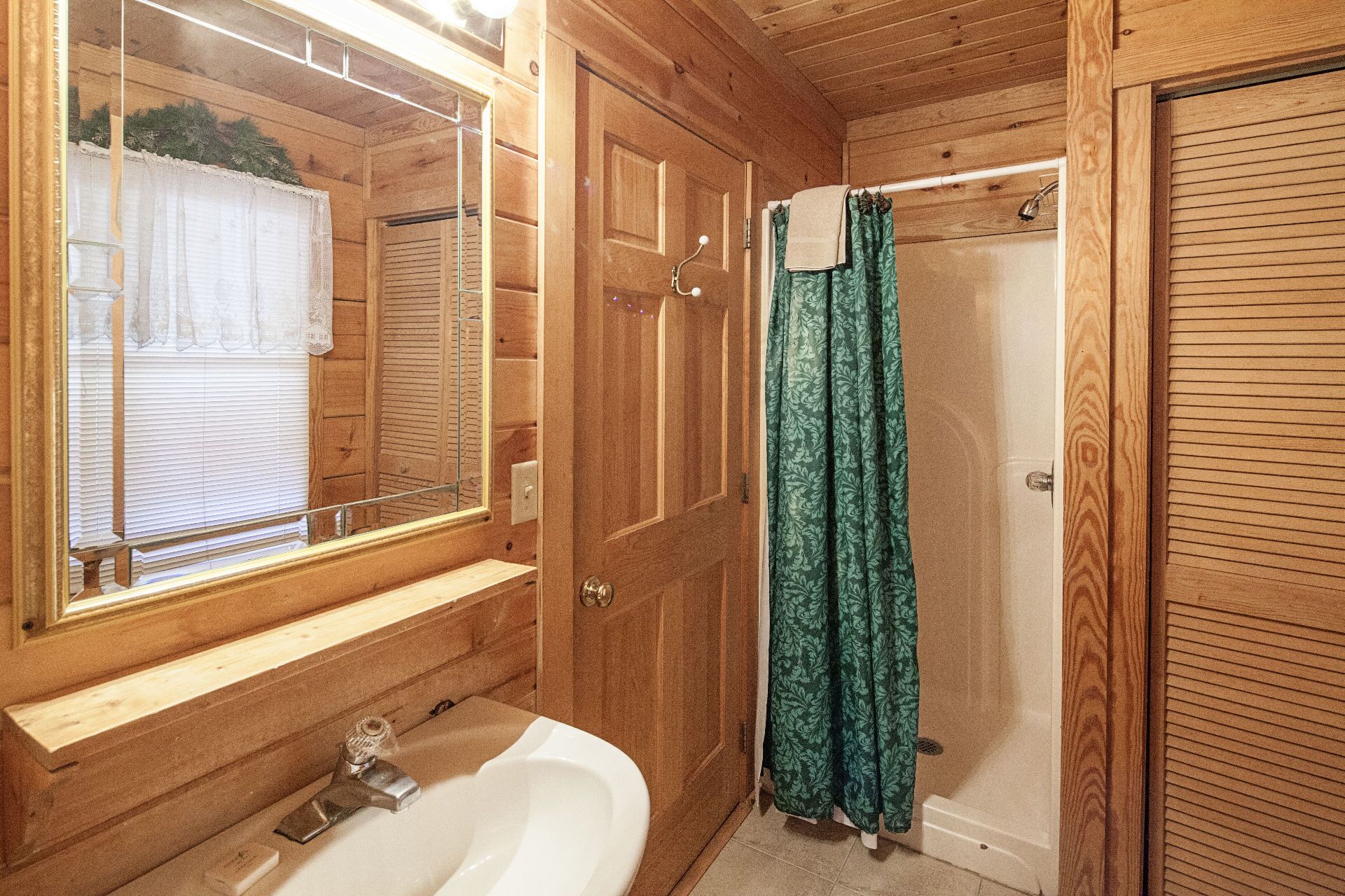 A wooden bathroom with a white sink, a mirror, a shower with a green patterned curtain, and wooden doors.