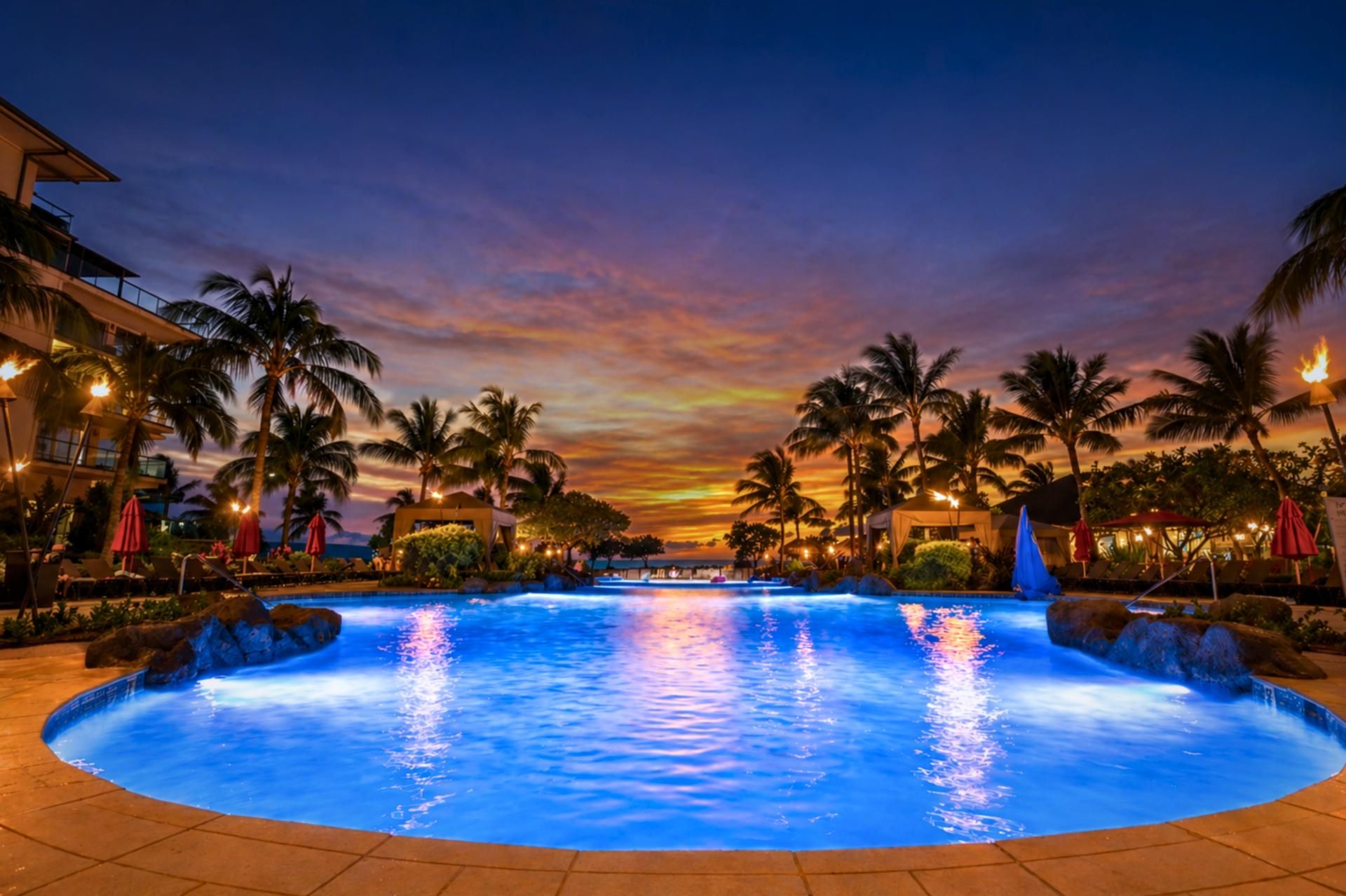 A swimming pool with a sunset view, surrounded by palm trees and hotel buildings.