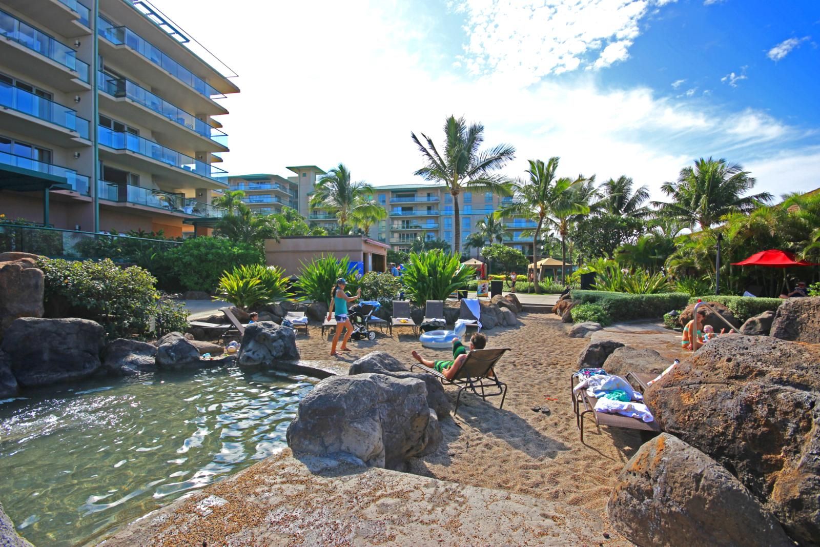 Een zwembadgedeelte van een resort met ligbedden, een fontein en palmbomen.