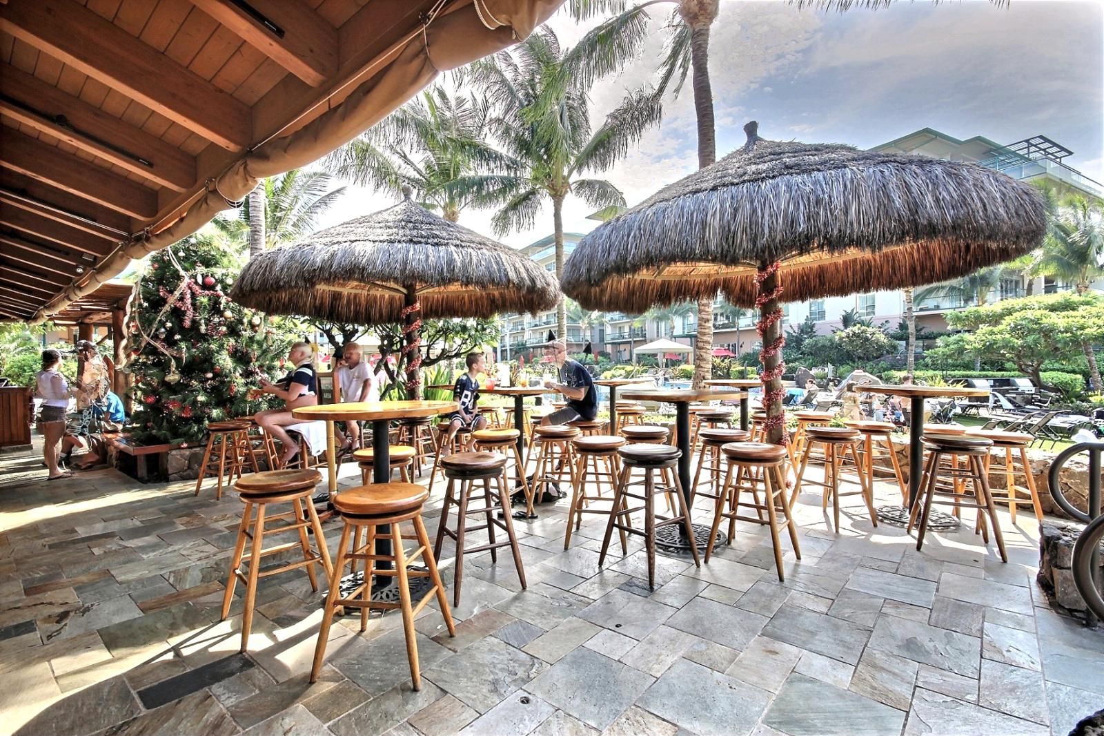 An outdoor seating area with thatched umbrellas, wooden tables, and chairs, surrounded by palm trees and a view of a resort.