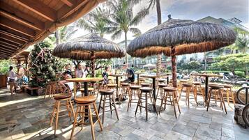 An outdoor seating area with thatched umbrellas, wooden tables, and chairs, surrounded by palm trees and a view of a resort.