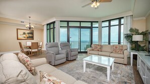 A spacious living room with a large window, a ceiling fan, and a dining area with chairs and a table. - Phoenix West - 24th Floor - 3BR (Orange Beach)