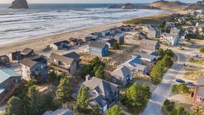 A coastal residential area with houses near the beach, a prominent rock formation, and a hilly landscape in the background.