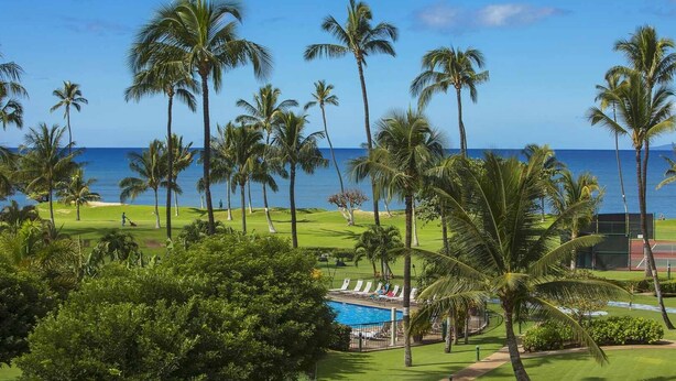 A golf course with a pool area, surrounded by palm trees and a view of the ocean. - Oceanfront Bliss • Ground Floor • Steps to Beach & Pool (Kihei)