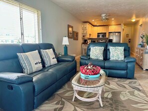 A living room with two blue leather sofas, a glass coffee table with decorative items, and a kitchen in the background.