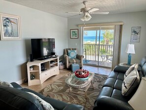 A living room with a television, a glass coffee table with a decorative bowl, a sofa, and a chair. There is a balcony with a view of the ocean.