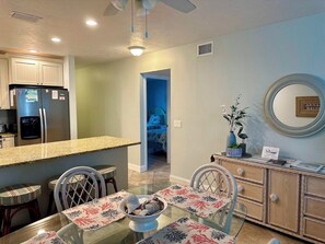 A dining area with a glass table, chairs, and a decorative centerpiece. There is a kitchen area with a refrigerator and cabinets. A mirror and a vase with flowers are visible on the wall.
