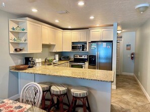 A kitchen with white cabinets, a granite countertop, a microwave, a stove, and a refrigerator. There are three bar stools and a small island with stools. - Island Beach Club 21 - Anna Maria Island - Condo 2 Bedroom / 2 Bath gulf front , maximum occupancy of 4 people. (Holmes Beach)