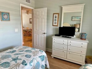 A bedroom with a bed, a dresser, a television, and a door leading to a dining area.
