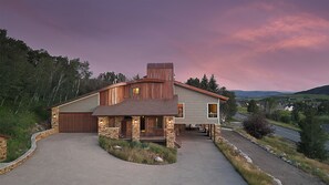 A two-story house with a stone facade, a wooden chimney, and a driveway leading to a gated entrance.
