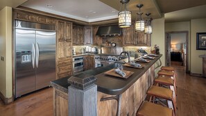 Une cuisine dotée de meubles en bois, d’un réfrigérateur en acier inoxydable, d’un grand îlot central avec des tabourets et d’un éclairage suspendu.