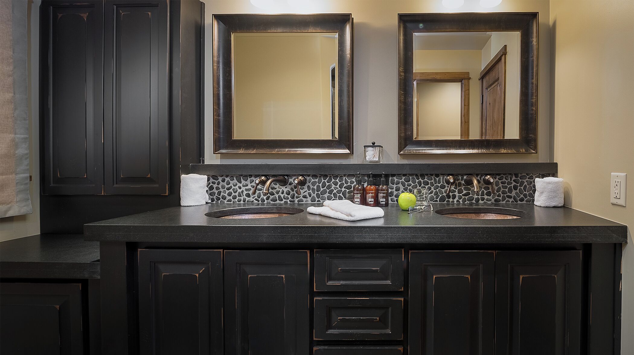 A bathroom with two sinks, dark cabinetry, a stone backsplash, and two mirrors.