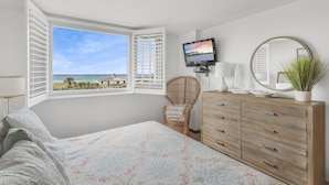 Ein Schlafzimmer mit großem Fenster, einem Bett, einem Schminktisch, einem Fernseher und einem Spiegel.