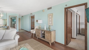 Ein geräumiges Wohnzimmer mit einem weißen Sofa, einem Holztisch und einem Schrank im Vintage-Stil. Ein Spiegel spiegelt einen Teil des Raumes wider, und eine Tür führt in einen weiteren Bereich.
