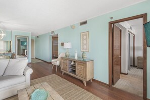 Un salon spacieux avec un canapé blanc, une table basse en bois et un meuble de style vintage. Un miroir reflète une partie de la pièce, et une porte donne accès à un autre espace.