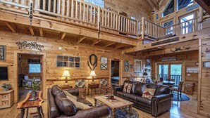Un espacioso interior de cabaña de madera con techo alto, un balcón y una zona de estar acogedora con muebles de cuero y chimenea.