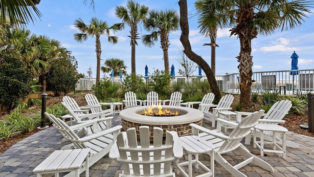 A circular fire pit surrounded by white Adirondack chairs on a paved patio with palm trees and a fence in the background.