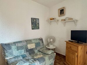 Um quarto com um sofá estampado, uma televisão sobre um armário de madeira, um ventilador e prateleiras fixadas na parede com objetos decorativos.