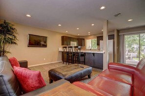 A living room with a sofa, ottoman, and a dining area with chairs and a kitchen in the background.