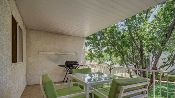 A covered outdoor area with a glass table and green chairs, a barbecue grill, and a view of trees and a white building.