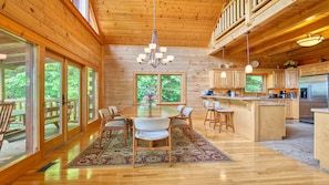 Cozy cabin with a high ceiling, dining area, kitchen, and fireplace, offering views of the surrounding greenery.