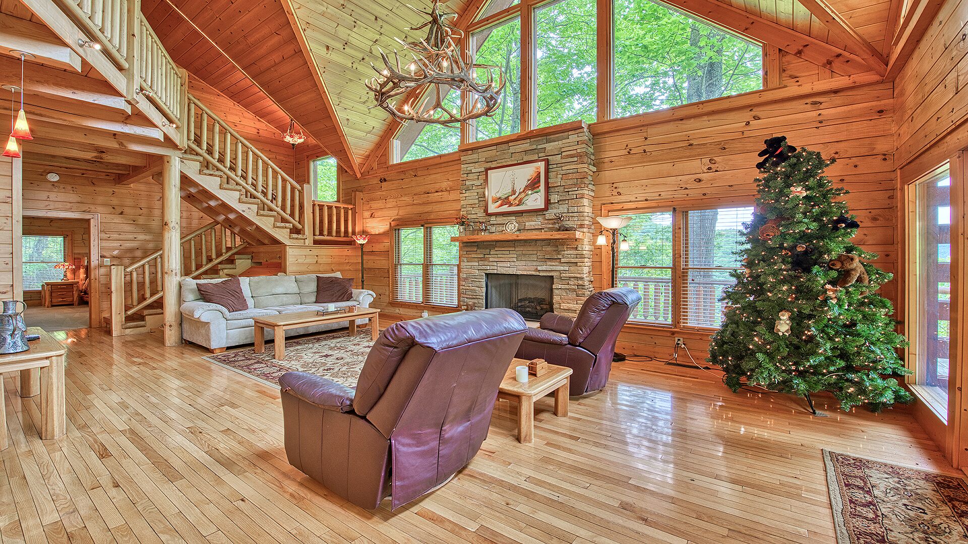 A spacious log cabin interior with a fireplace, a Christmas tree, and a staircase leading to an upper level.