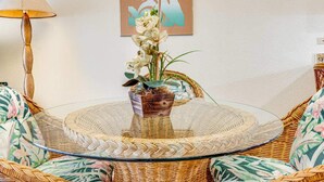 Una mesa redonda de cristal con una base trenzada, rodeada por sillas de mimbre con tapicería floral. Una orquídea en maceta está colocada sobre la mesa.