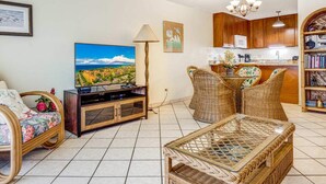 Una sala de estar con televisor, mobiliario de mimbre, piso de baldosas y una cocina al fondo.