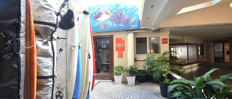 Un vestíbulo de hotel con tablas de surf apoyadas en una pared, un cartel de consigna de equipaje y plantas en macetas.