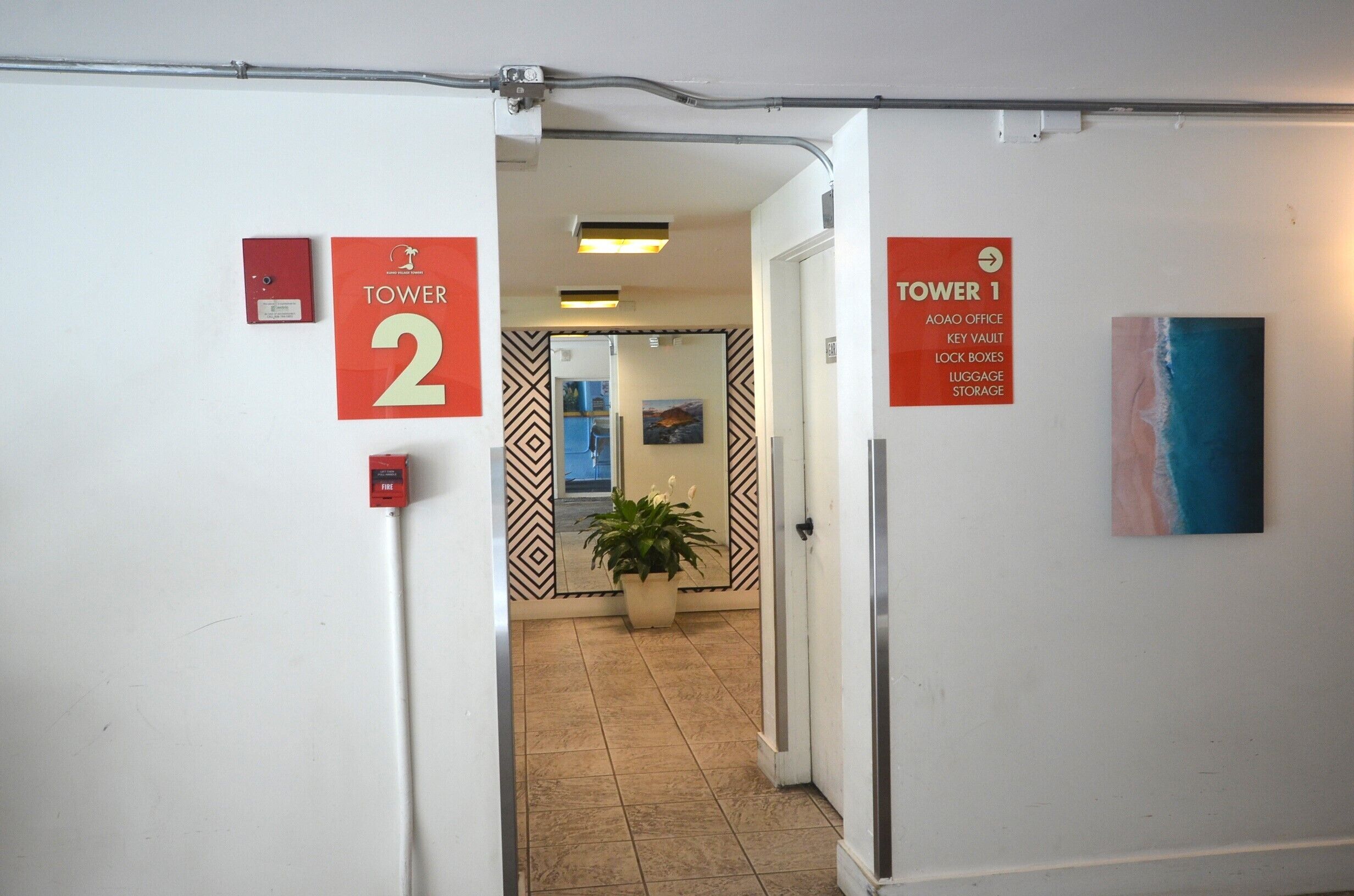 A hallway with signs indicating 'TOWER 2' and 'TOWER 1' with amenities listed such as 'AOAO OFFICE,' 'KEY VAULT,' 'LOCK BOXES,' 'LUGGAGE STORAGE.'