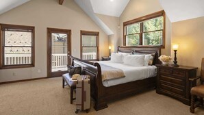 A bedroom with a large bed, a dresser, a bench, and a suitcase. There are windows with blinds and a view of the outdoors.