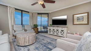 Ein Wohnzimmer mit rundem Couchtisch, zwei Sesseln, einer Couch, einem Fernseher auf einer weißen Kommode und einem Balkon mit Blick.