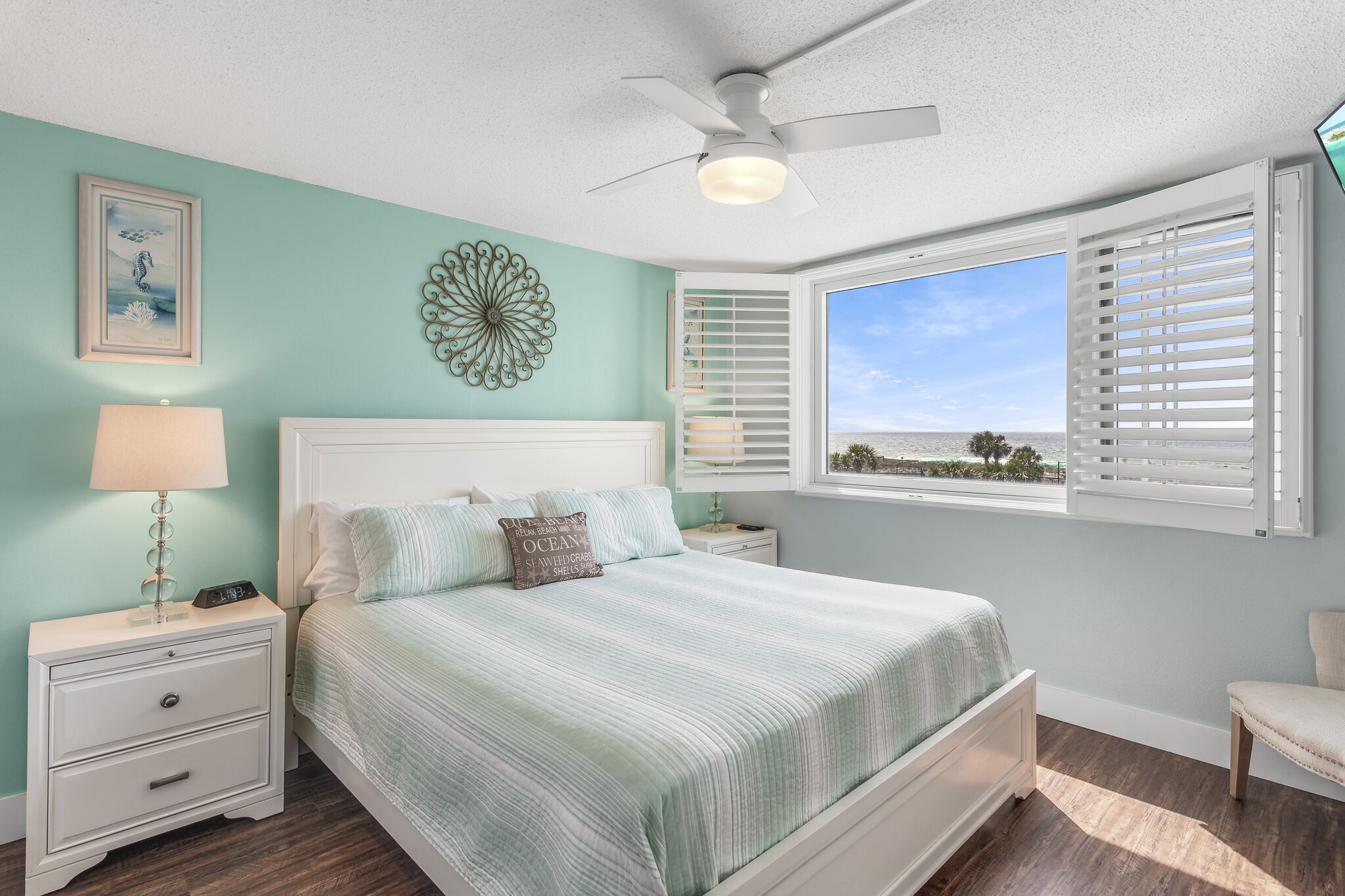 A bedroom with a bed, nightstands, a ceiling fan, a window with blinds, and a view of the ocean.