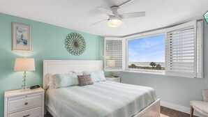 Ein Schlafzimmer mit einem Bett, Nachttischen, einem Deckenventilator, einem Fenster mit Jalousien und Meerblick.