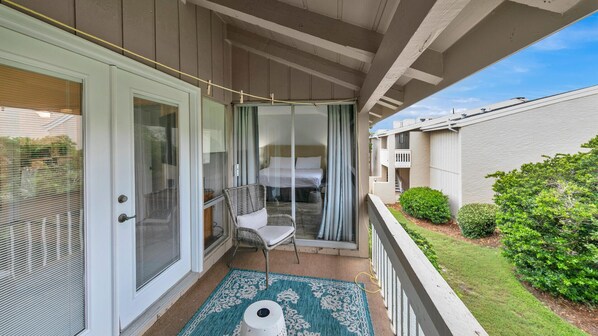 A balcony with a wicker chair, a small rug, and a view of a bedroom with a bed and curtains. - Chateau La Mer 9D | Private Beach | FREE Seasonal Activities (Destin)