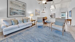 Amplia sala de estar con un sofá de color claro, dos sillones, una mesa de centro y un tapete azul. Se observan un ventilador de techo y una araña.