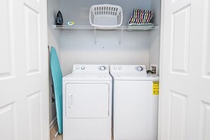 A laundry room with two white washing machines, a drying rack, and a shelf with laundry supplies.