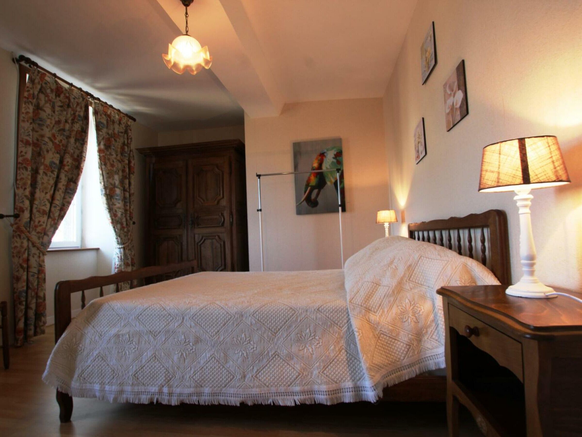 Un dormitorio con cama, mesita de noche, lámpara, ventana con cortinas, puerta y una pared con un cuadro.