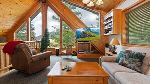 Ein gemütliches Zimmer mit Holzbalkendecke, großen Fenstern, einem Fernseher, einem Sofa, einem Couchtisch und einem Bücherregal.