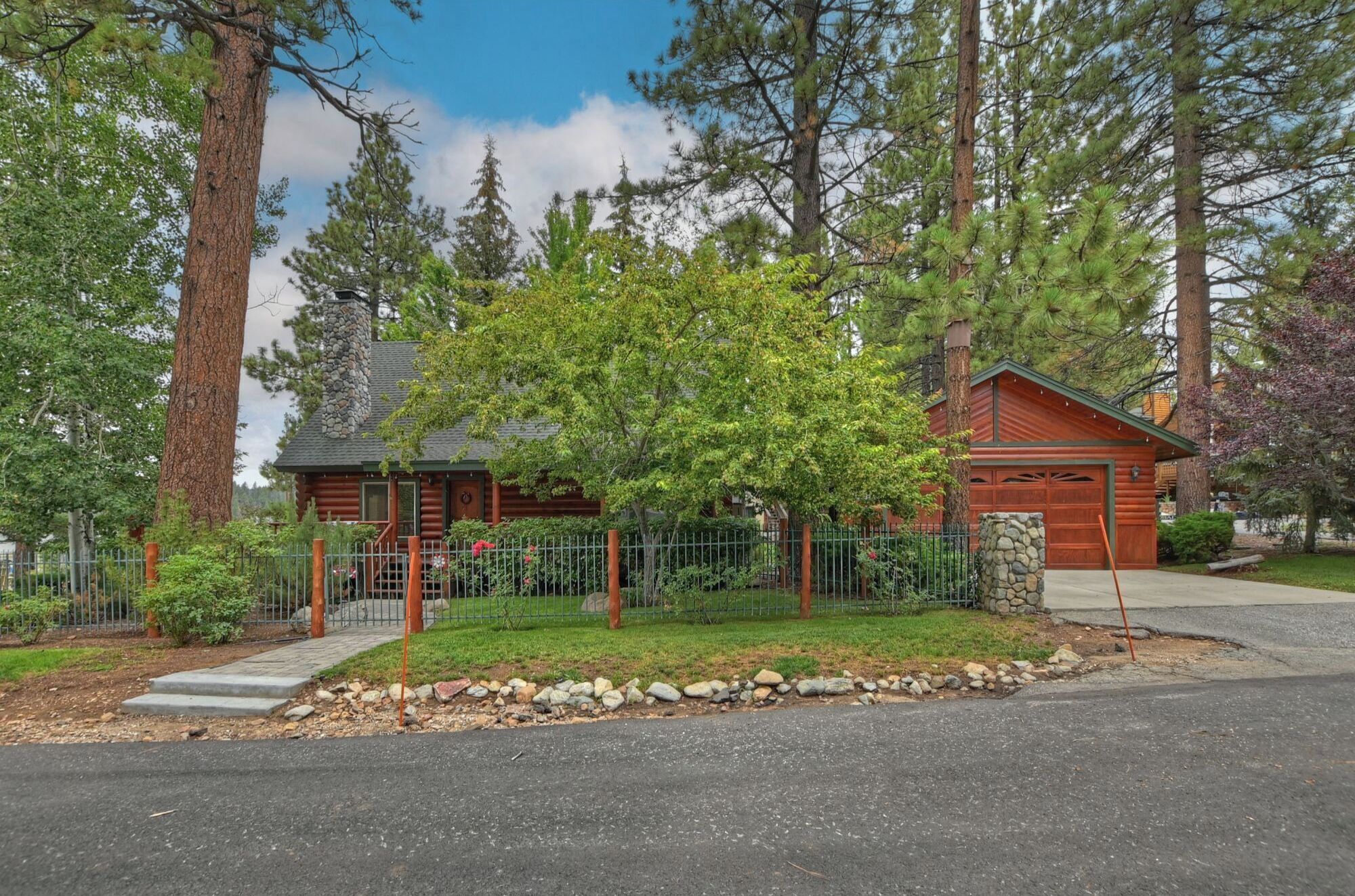 A log cabin with a stone chimney, surrounded by a stone wall and a metal fence.